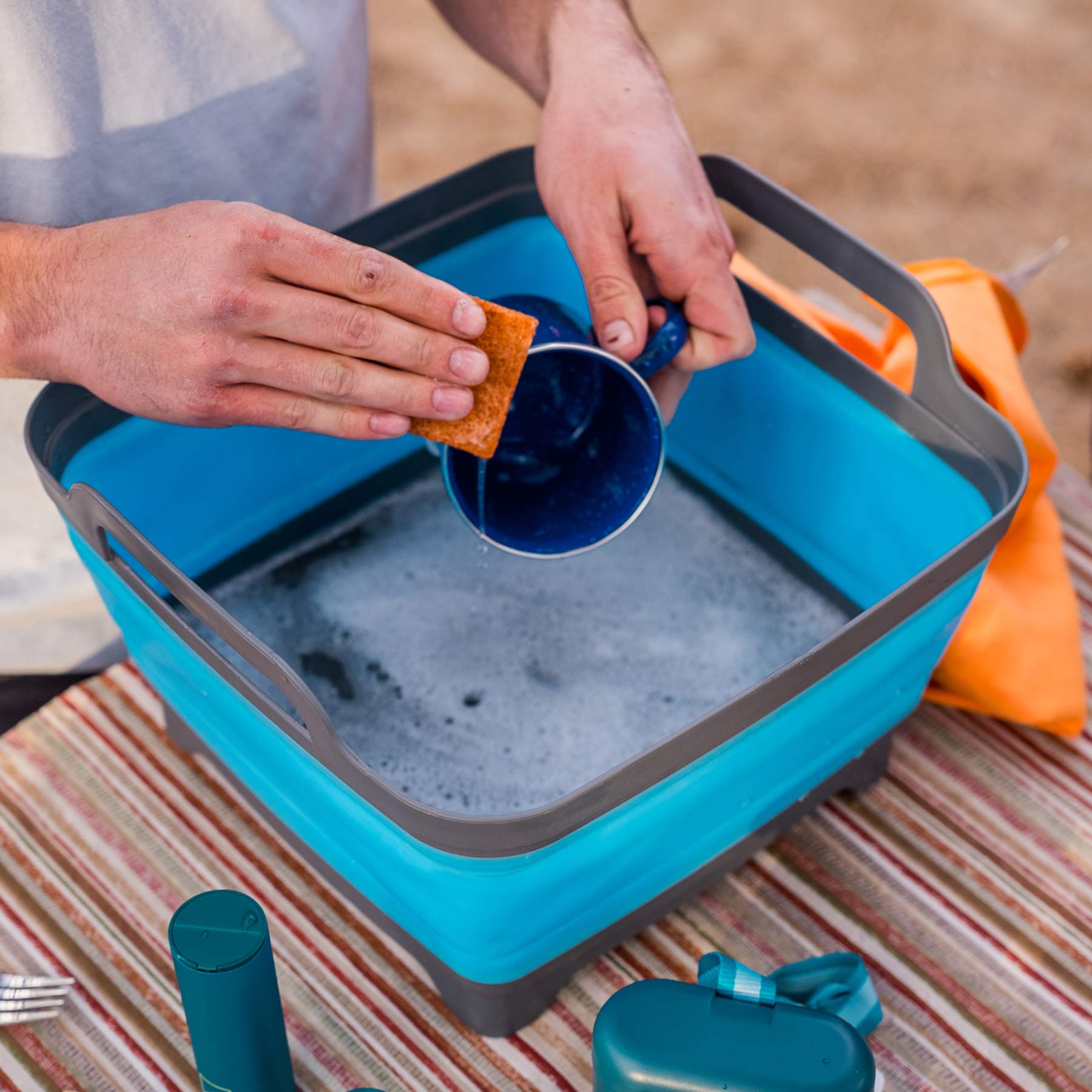 A person using the coconut scrubber at a campsite to wash a cup in a collapsible sink.