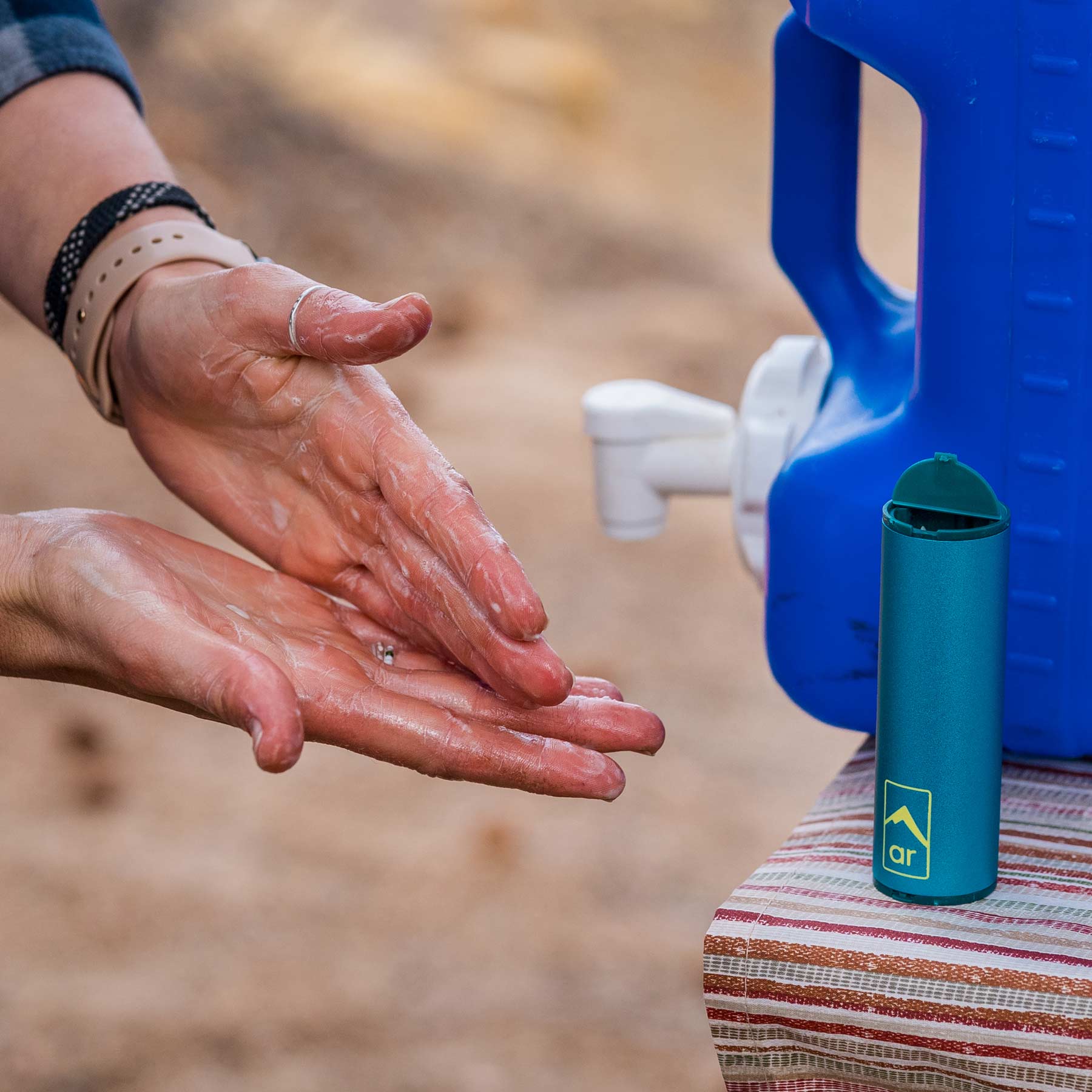 Image shows a person washing their hand in the wild lathering up using the soap flakes. It also shows the clean capsule on a table next to a jug of water.
