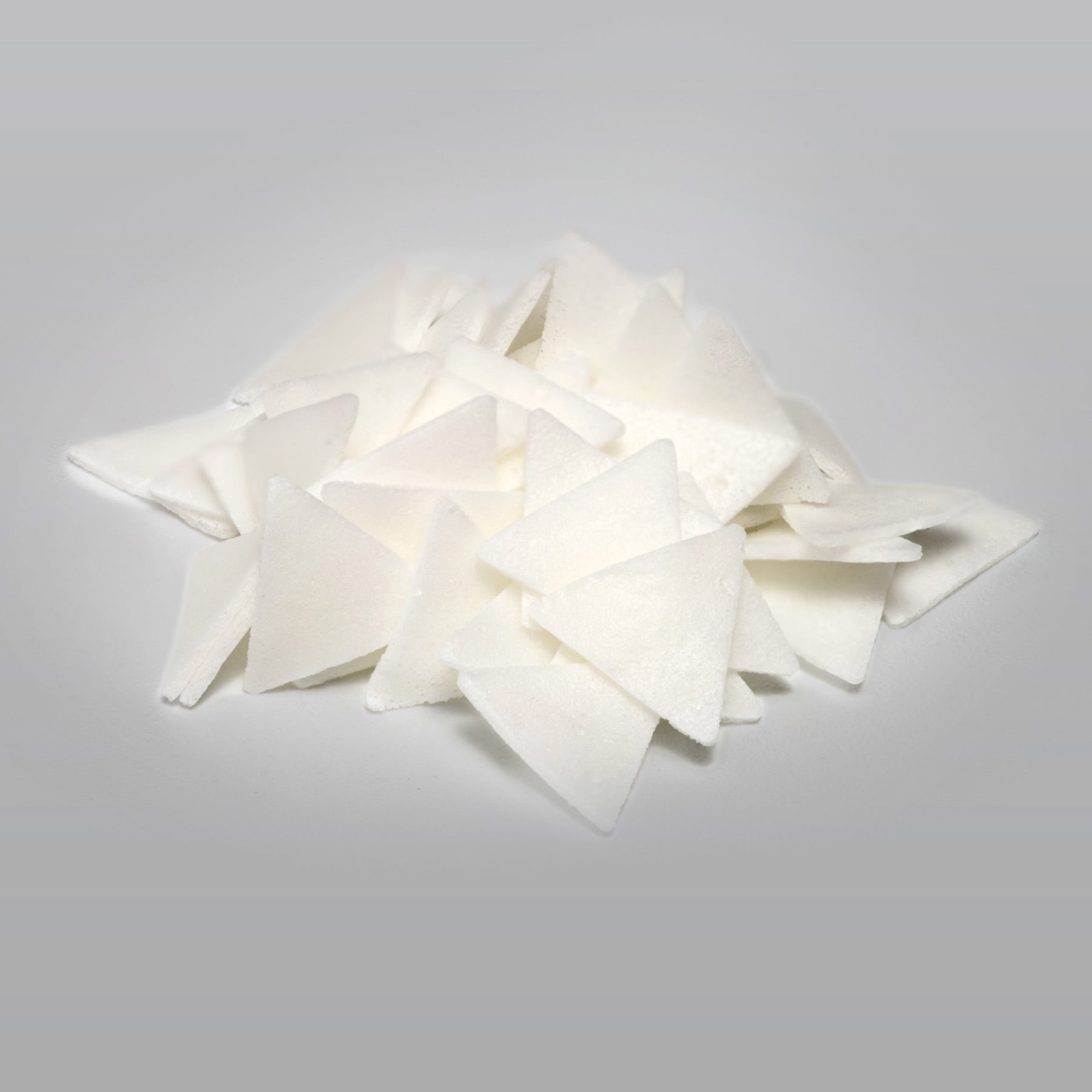 A pile of white triangle shaped AR soap flakes on a white/grayish background.