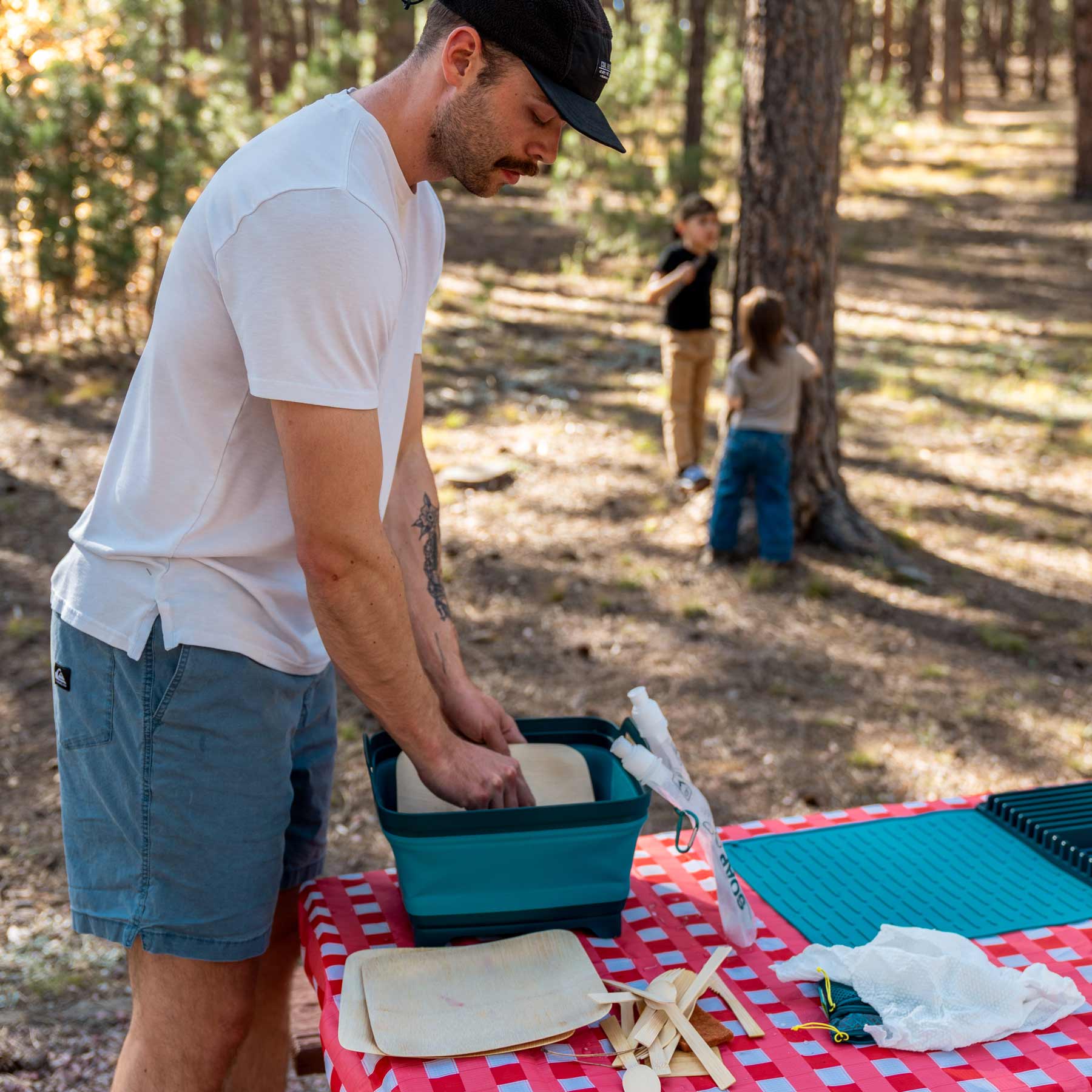 A man is washing some bamboo dishes in a collapsible sink at a campsite.
