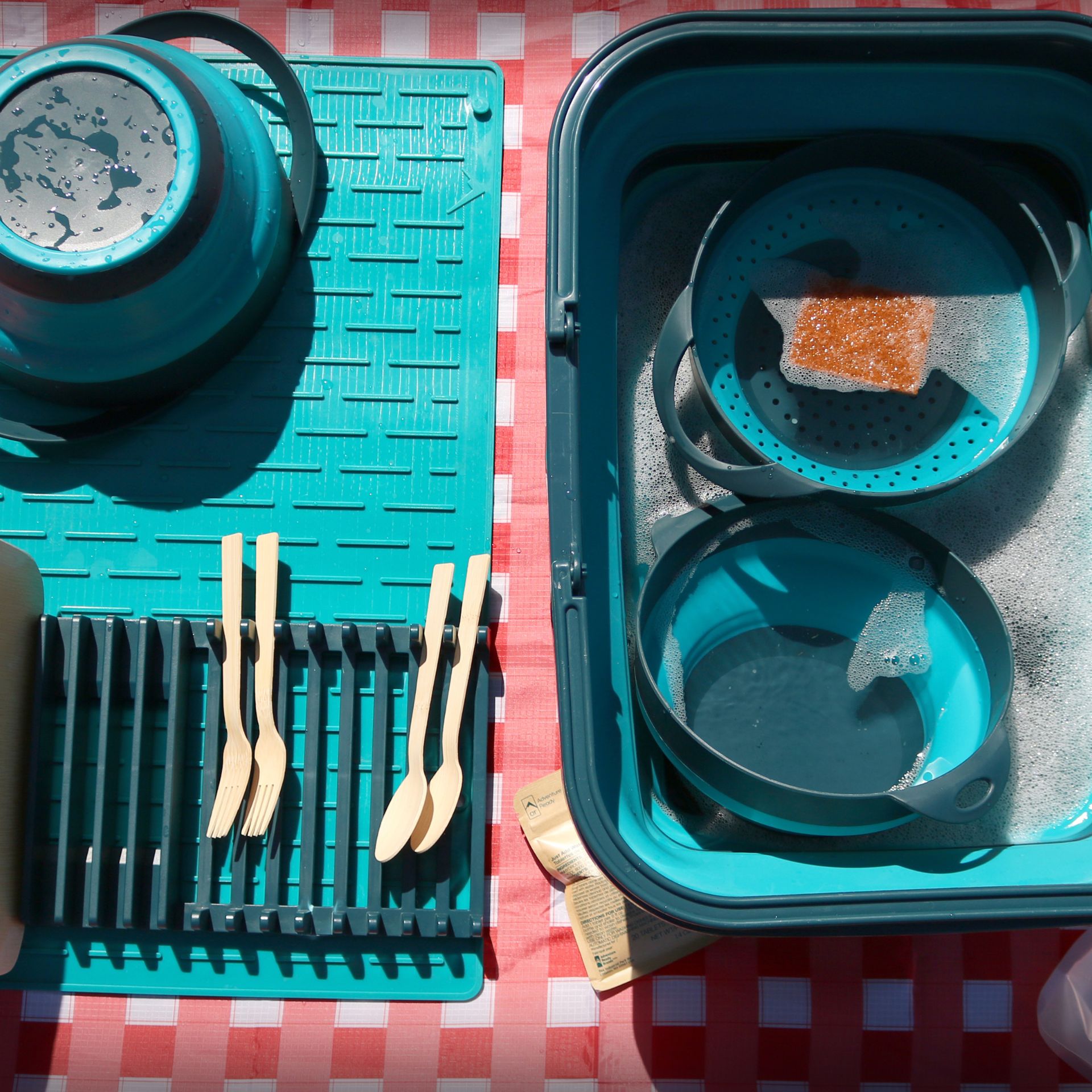 On the left showcases the AR Roll-Up Dish Drying Station. On the mat is a wet bowl from the AR Collapsible Bowl and Strainer Set. The dish rack is also sitting on the mat and is drying the AR Bamboo Dining Set's forks, spoons, and plate. To the right is the AR Collapsible Sink 16L filled with soapy water. Soaking in the water are a bowl and strainer from the Bowl and Strainer set. Floating on top of the water is the AR Coconut Scrubber. All of this is set on top of the AR Picnic Tablecloth, Red Gingham.