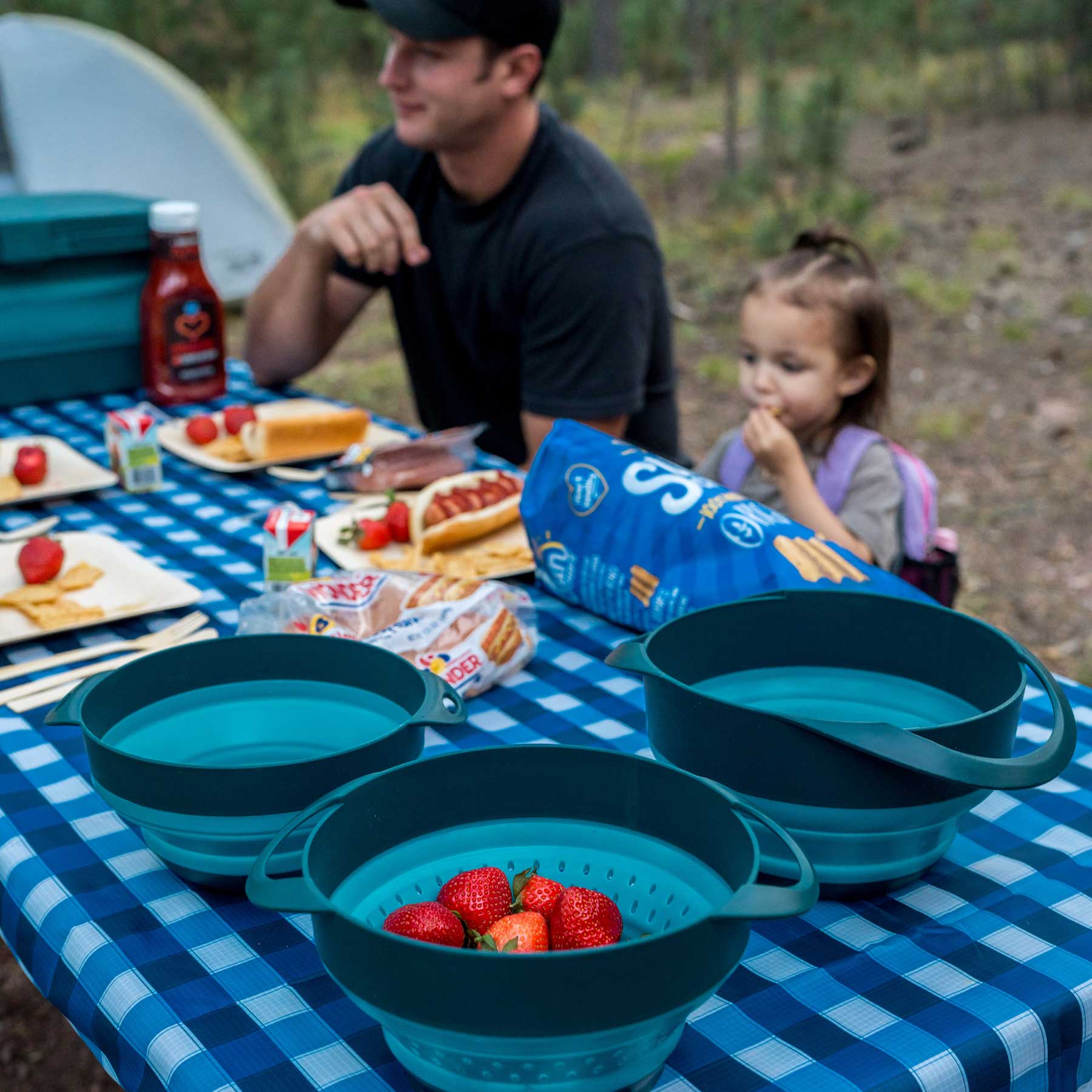 The image shows a man and a child in the back eating from their AR Bamboo Dining Set. In the foreground shows the Flat Pack Collapsible Bowls and Strainer. The strainer has strawberries in them. The setting is at a campsite in the woods.