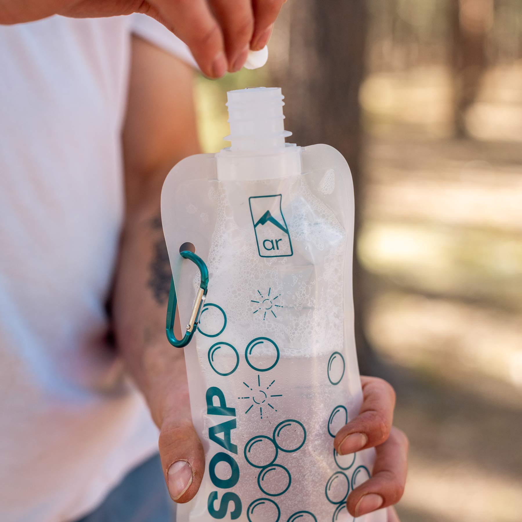 Image shows a person holding the holding the "Soap" water bottle from the Adventure REady Just-Ass-Water Dishwashing Kit in one hand and ready to drop a dishwashing soap tablet into it with the other hand. The water in the bottle is already soapy.