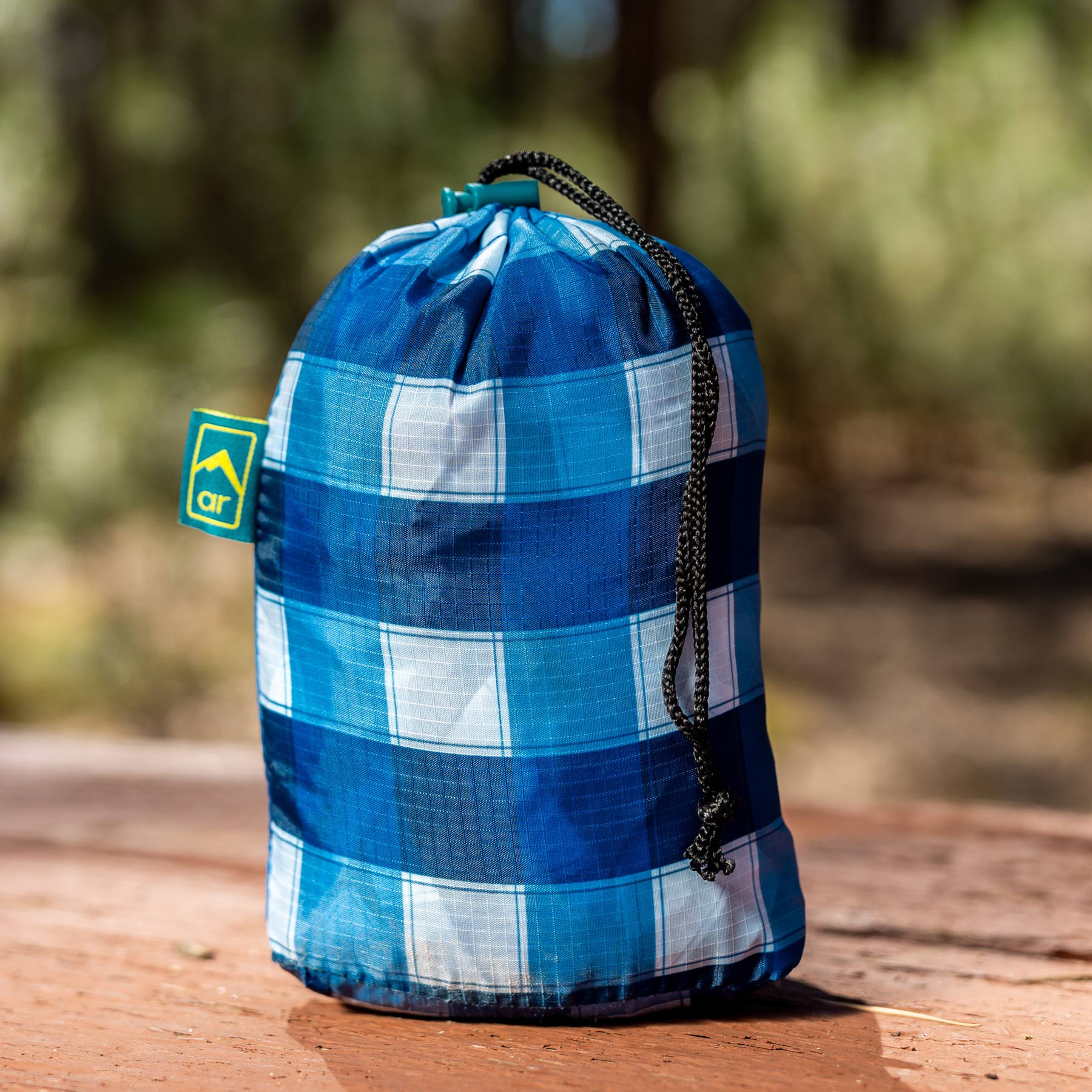 The Adventure Ready Picnic Tablecloth - Blue Gingham in stuff sack on a table outdoor.