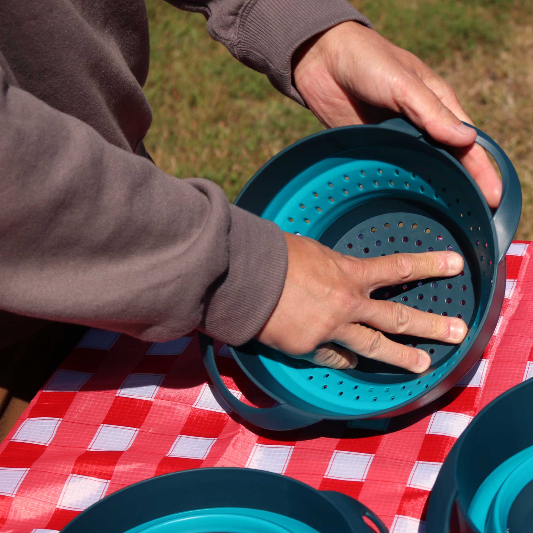 A person pushing to expand the Flat Pack Strainer on top of a picnic table covered with the AR Picnic Tablecloth.