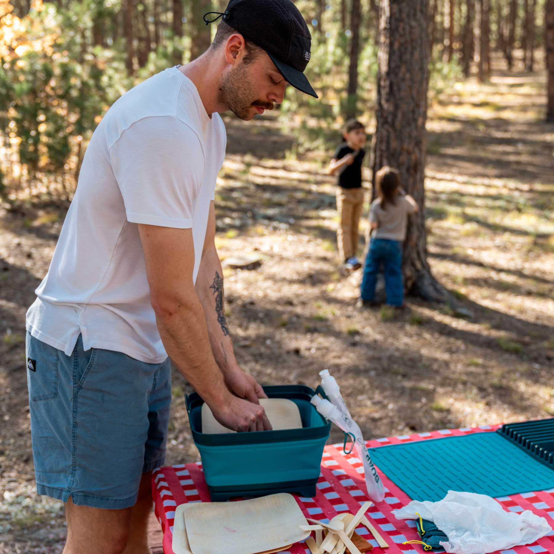 A man is washing some bamboo dishes in a collapsible sink at a campsite. 