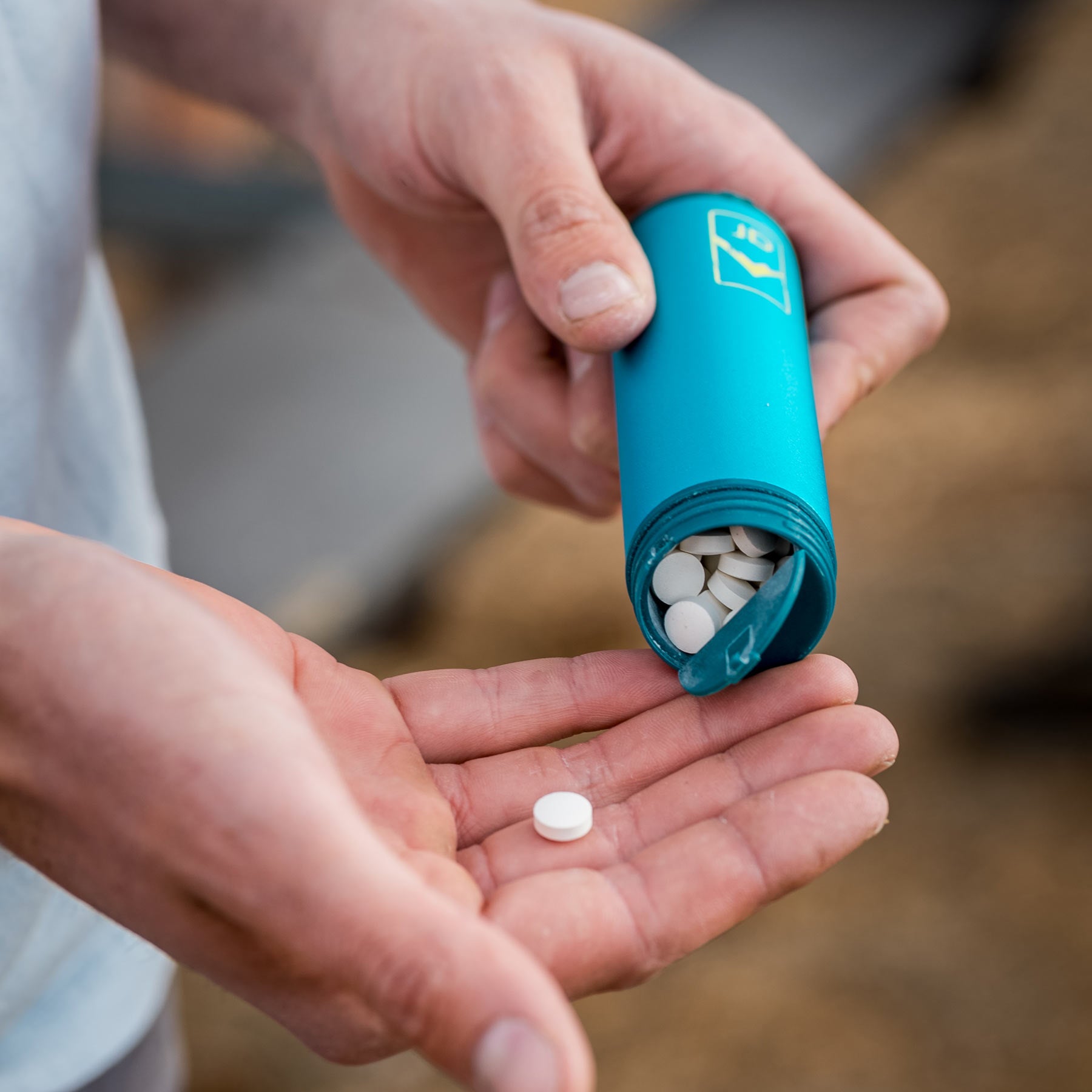 The image shows in hand, in an outdoor environment, the toothcare capsule open, dispensing a toothpaste tablet into the user's hand. The tablet is white and the about the size of a breath mint.