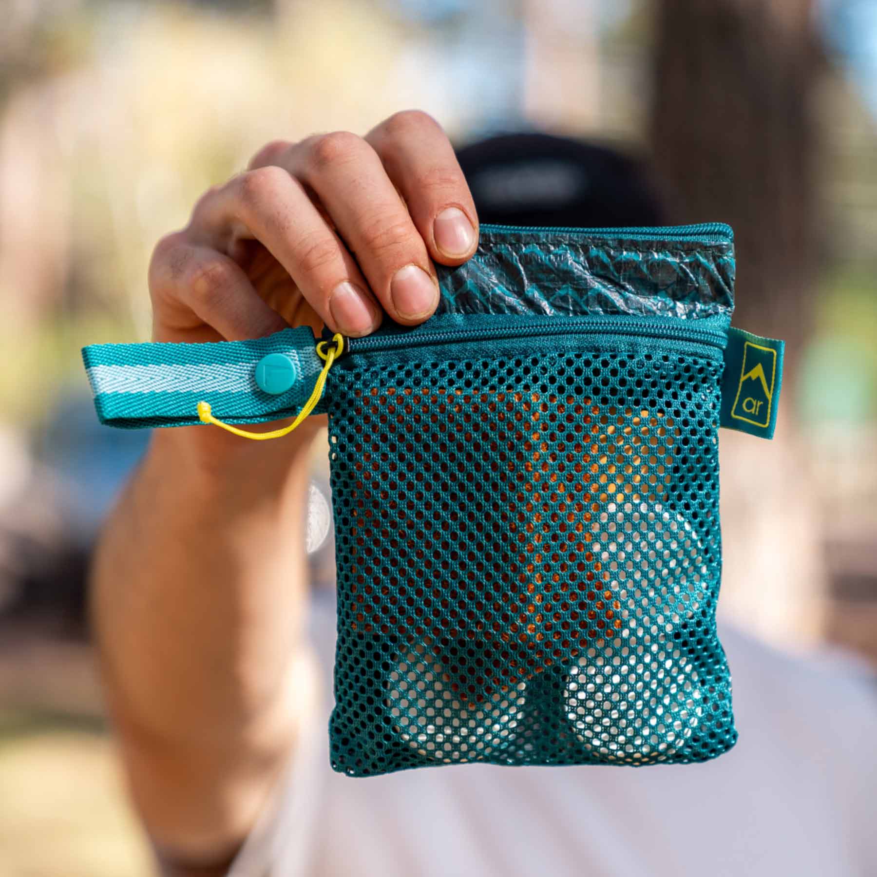 Image is a person holding the Adventure Ready Scrub & Dry Clean-Up Kit in front of them. This shows the bag holding the contents. The mesh side shows visibly the coconut scrubber, bamboo scraper, and compressed towels.